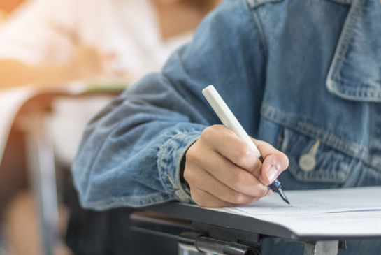 A students hand is seen writing on papers on a desk in a classroom. 