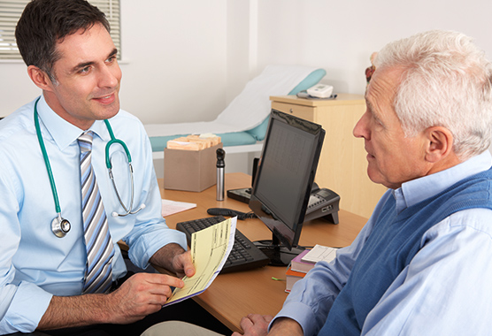 Male health care provider talking to older male patient at a desk in a clinic.