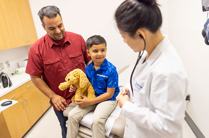 Young boy sitting on an exam table holding a teddy bear next to his dad and female health care provider