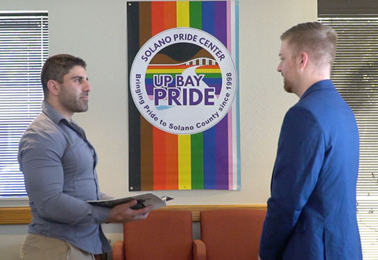 Two men facing each other conversing in front of the colorful rainbow flag logo belonging to Solano Pride Center in Fairfield