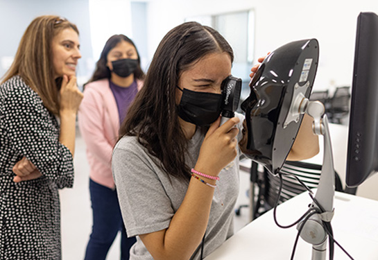 Girl wearing mask holds otoscope up to her eye and looks into manikin face while two women observe standing behind her