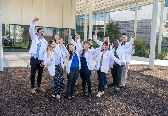 Group of students wearing white coats stand side-by-side in a group with raised arms in celebration