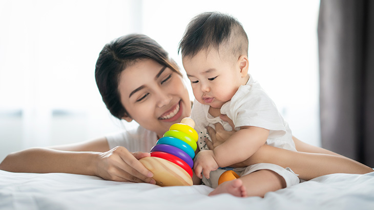 A smiling woman with dark hair pulled back kneels beside a bed, her arm around an infant sitting on the bed who is playing with a colorful wooden stacking toy.