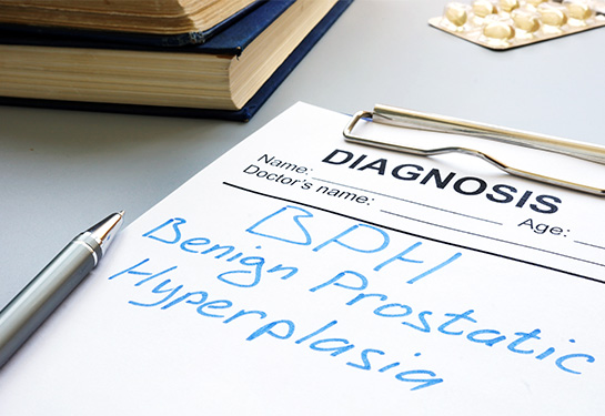 A clipboard holding white paper and the terms “diagnosis, Benign prostatic hyperplasia” with a silver pen, packet of yellow pills and two large books