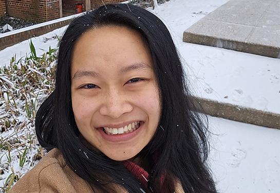 A woman with dark hair past her shoulders smiles in front of a red brick building near snow on the ground