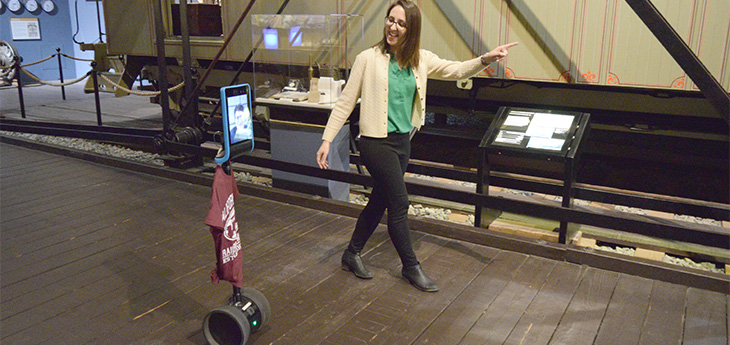 Woman walks next to two-wheeled robot with a video screen in museum.  