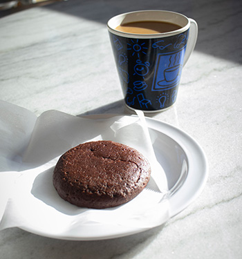 A chocolate cookie on a white plate sits next to a blue and black coffee mug on a marble tabletop. 
