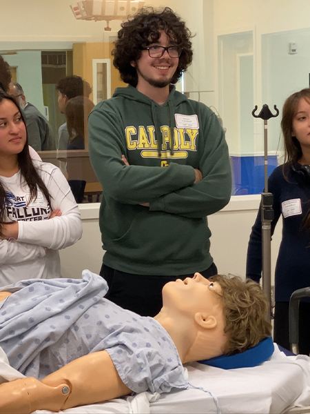 A teenage boy in a green sweatshirt smiles and stands beside a training manikin as he learns life-saving techniques