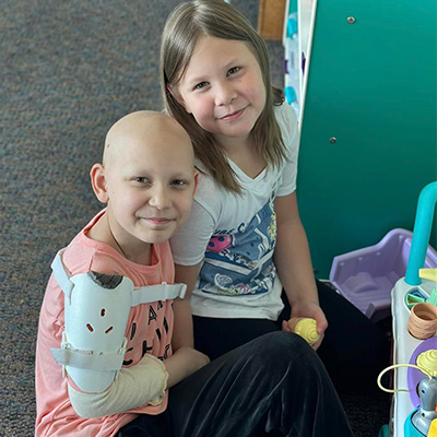A smiling child with an arm brace who has lost her hair and another child play in the playroom.