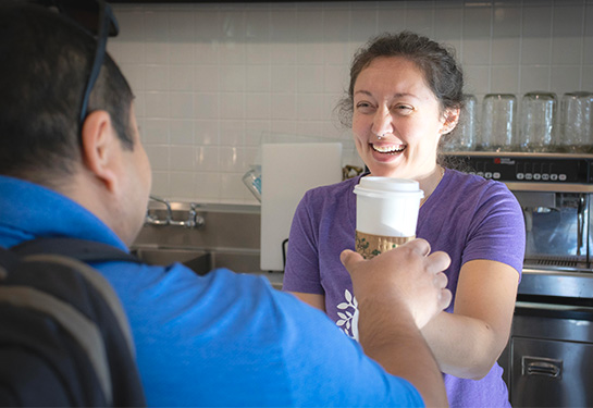 A female café employee in a purple T-shirt hands a travel coffee cup to a customer while smiling broadly. 