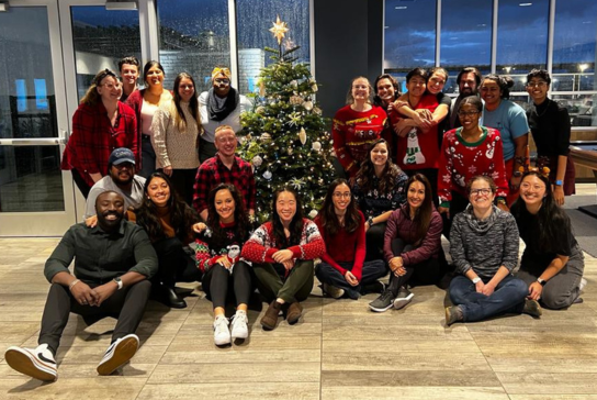 A large group of family medicine residents, some wearing holiday sweaters, smile around a holiday tree.