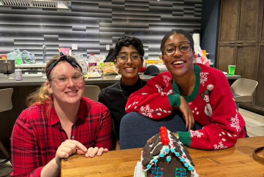 Three women, family medicine residents, smile in front of a gingerbread house.