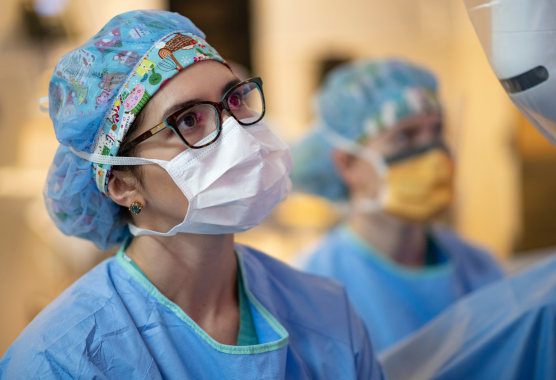 A woman wearing a mask and scrubs with glasses looks with focus to her left.