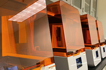 Four 3D printers with orange covers sitting on a table in a row.