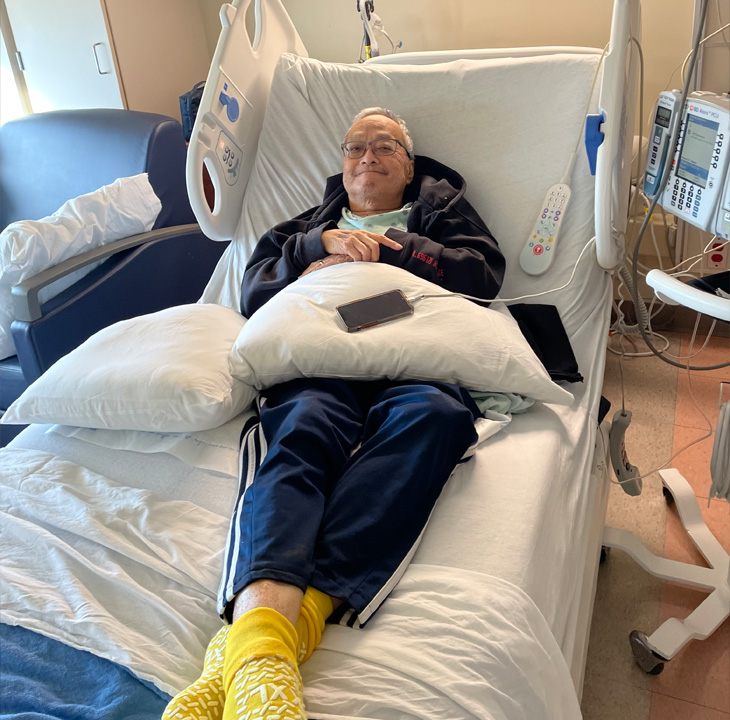 An older man wearing glasses, navy track pants, yellow socks and a navy jacket lies in hospital bed with arms crossed and smiling