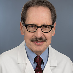 A man with brown hair and wearing glasses and a white physician’s coat,  smiles