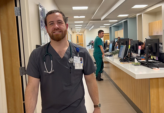 A man with dark hair and beard, wearing gray scrubs, walks down hallway of a medical clinic 