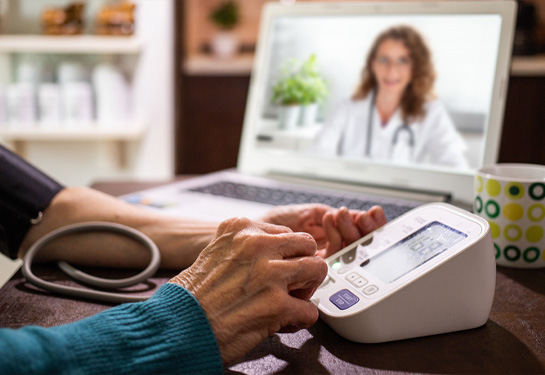Close up of elderly hands using blood pressure machine at home while physician looks on in the background from a computer laptop screen