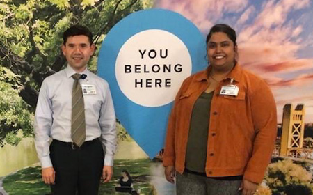 Two smiling persons standing in front of a huge poster with a “YOU BELONG HERE” message. The person on the left is wearing a grey shirt, a green stripped necktie, and black pants. The one on the right is wearing an orange jacket on top of a dark colored short.