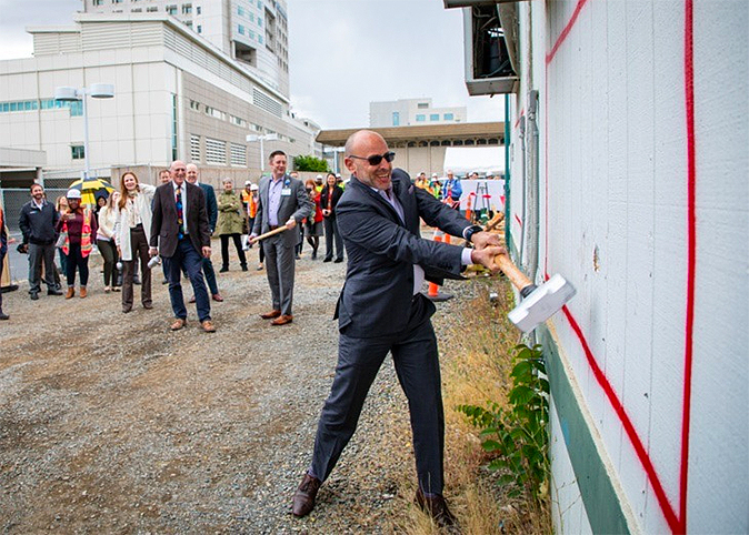 man in a suit swinging a sledgehammer into a wall at a groundbreaking ceremony