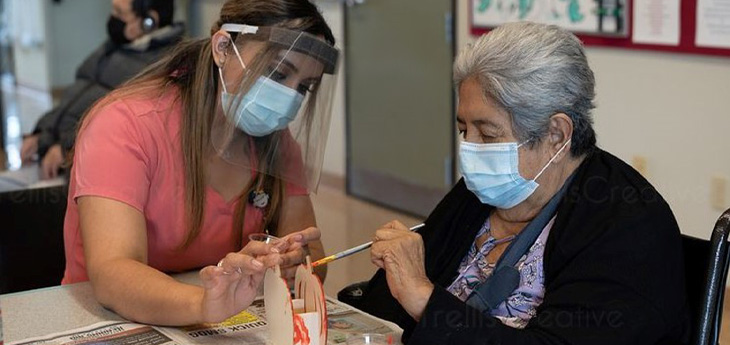 woman wearing mask and face shield helps older woman wearing mask paint a ceramic pumpking