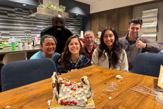 Five family medicine residents smile and laugh in front of a broken gingerbread house.