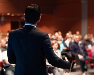 image of a male speaker in the foreground addressing an audience in the background